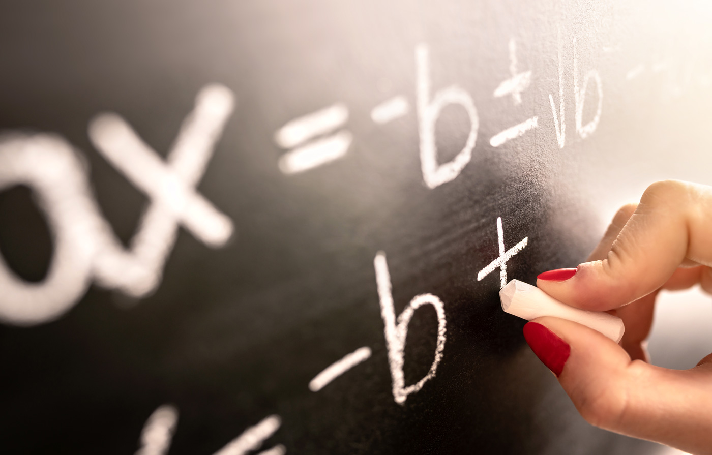 A person with red nails writes algebra equations on a chalkboard with a white piece of chalk