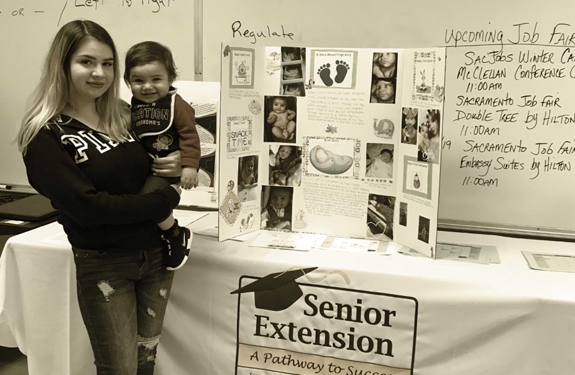 a smiling young woman stands in front of a "Senior Extension" event table holding a smiling toddler