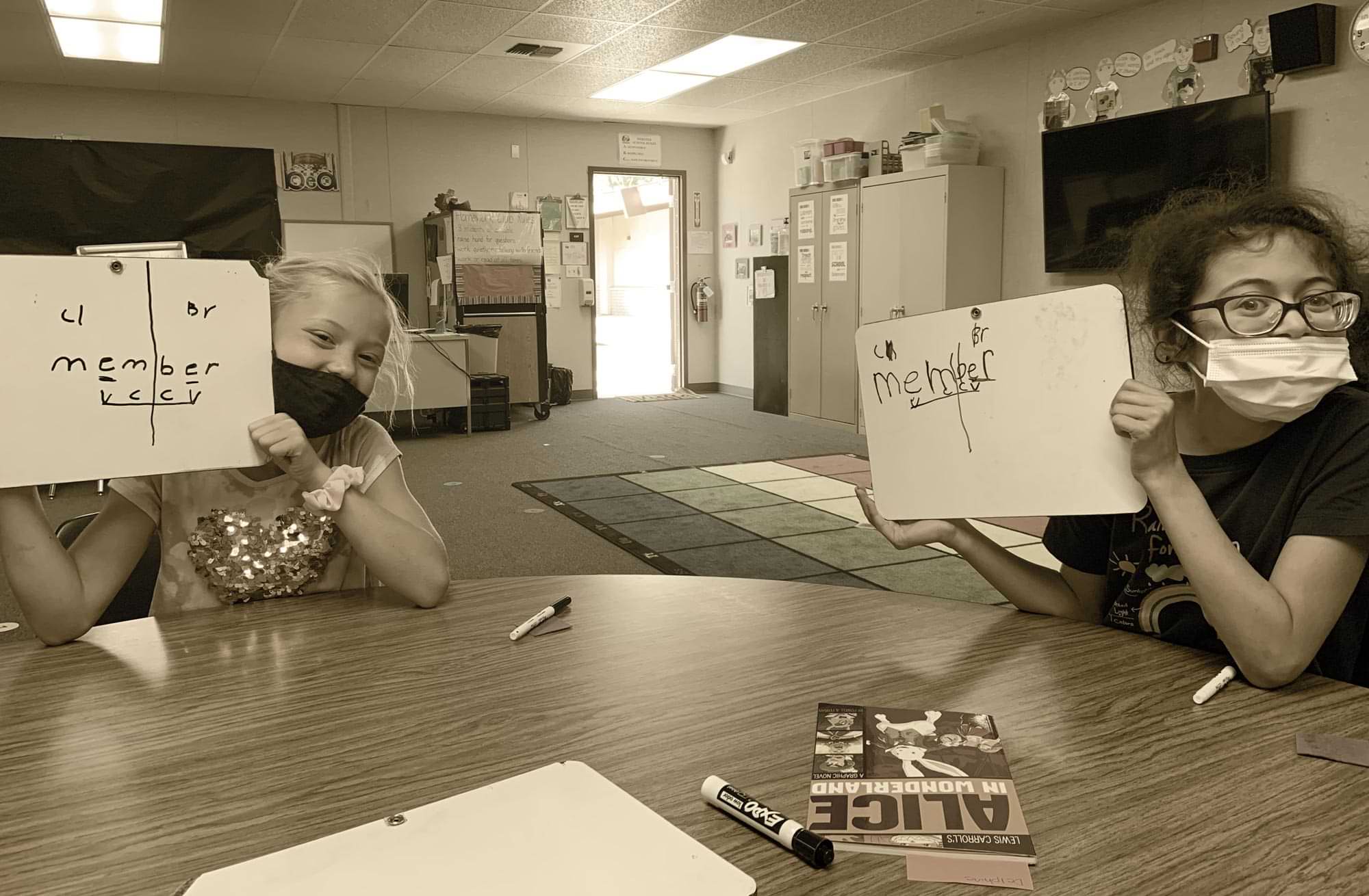 two masked elementary aged students study reading with the help of white boards in a classroom