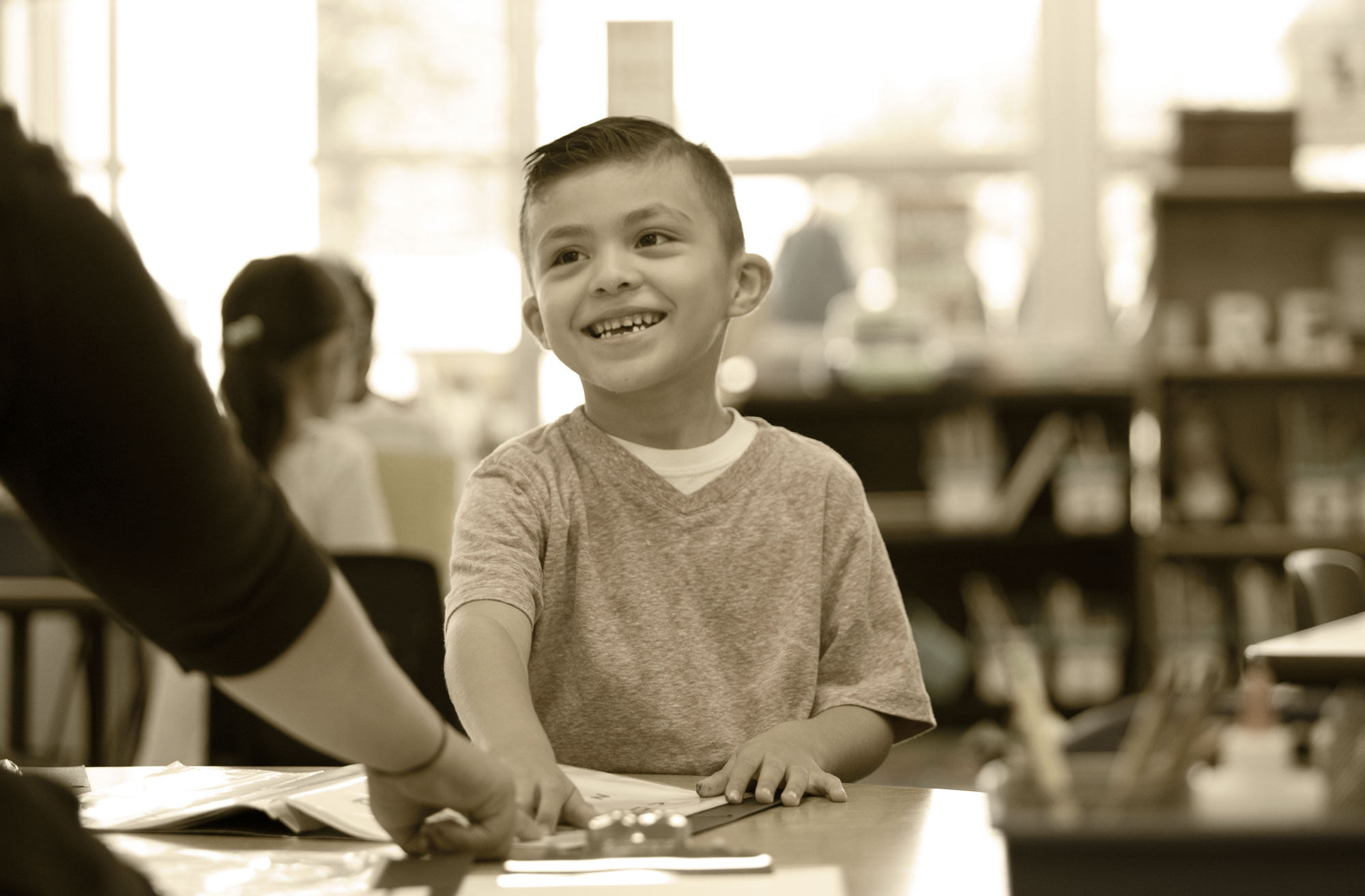 a young boy smiles sitting at a desk studying with an adult