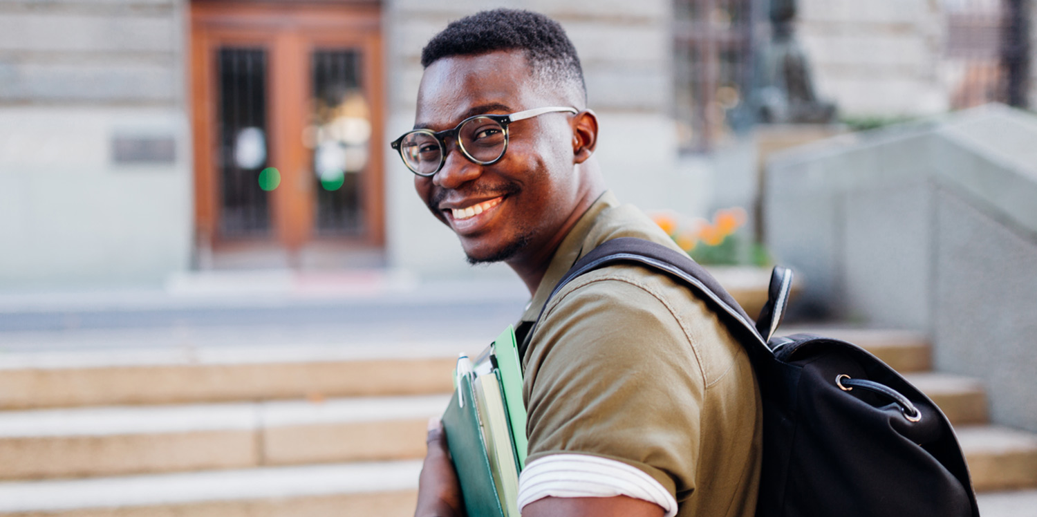 a black male student wearing glasses and a backpack and holding books smiles while walking toward a building