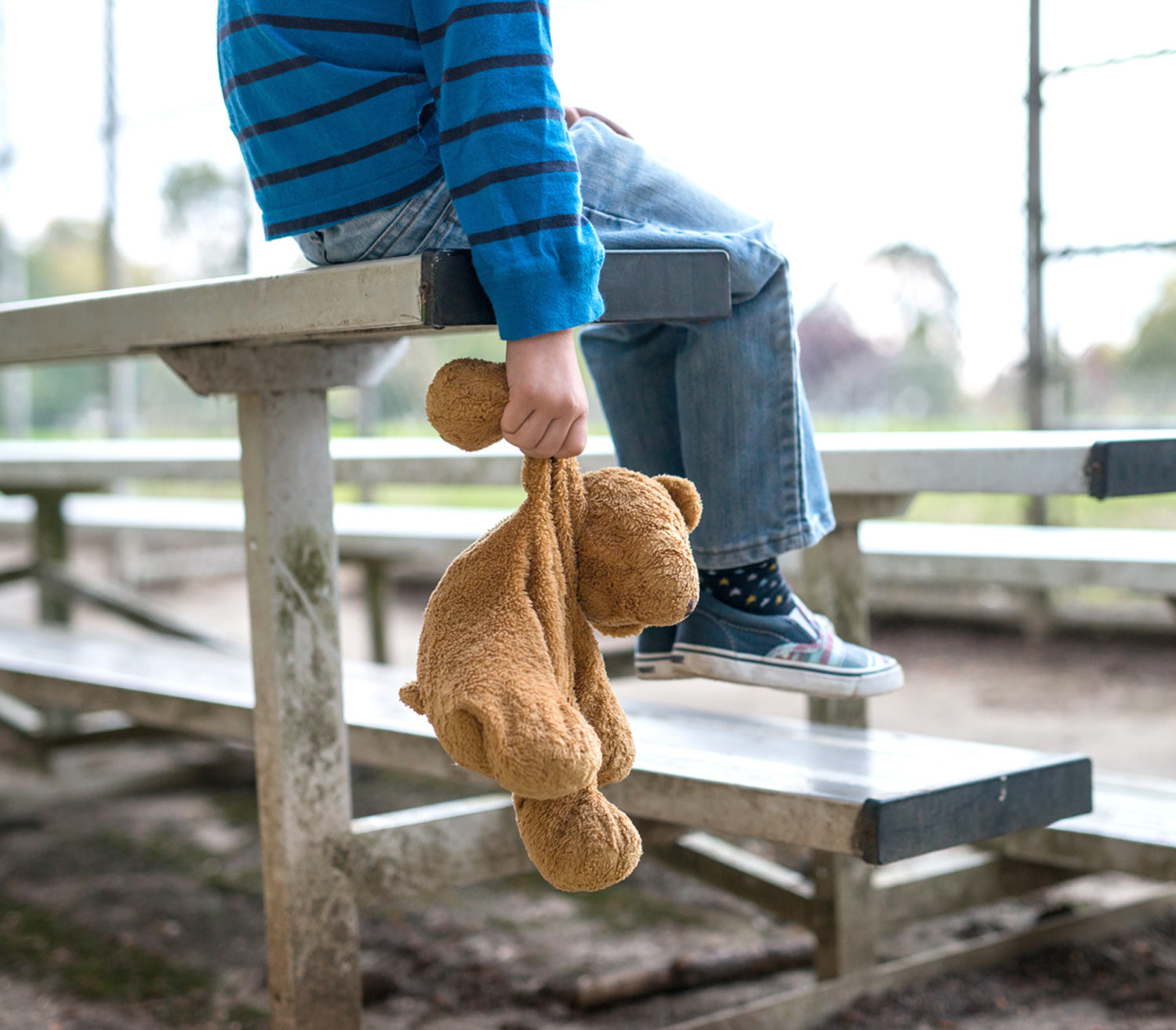 a child sits alone on a bleacher, holding an old teddy bear by the arm