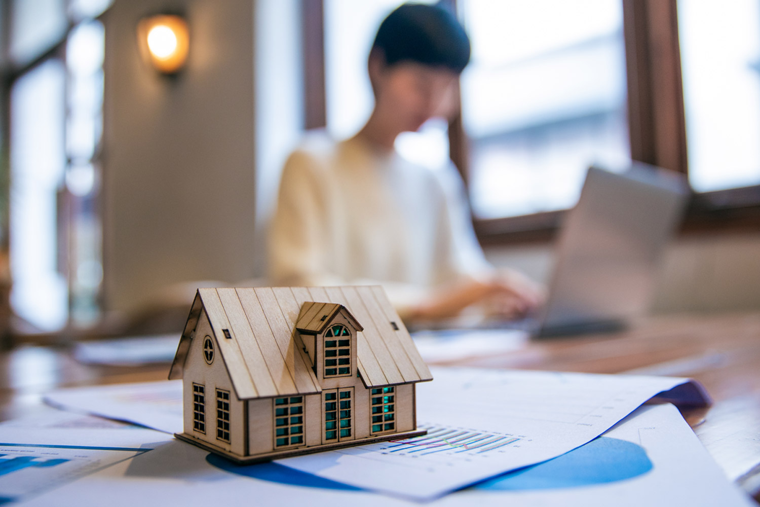 a small model house sits on a table with paper in the foreground while a person works on a laptop out of focus in the background