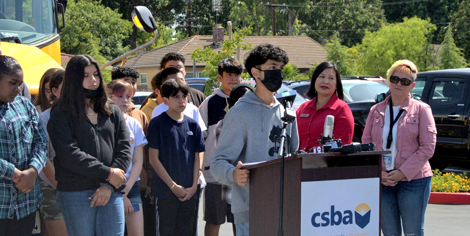 a teenager wearing a face mask speaks at a podium with a crowd of others behind him