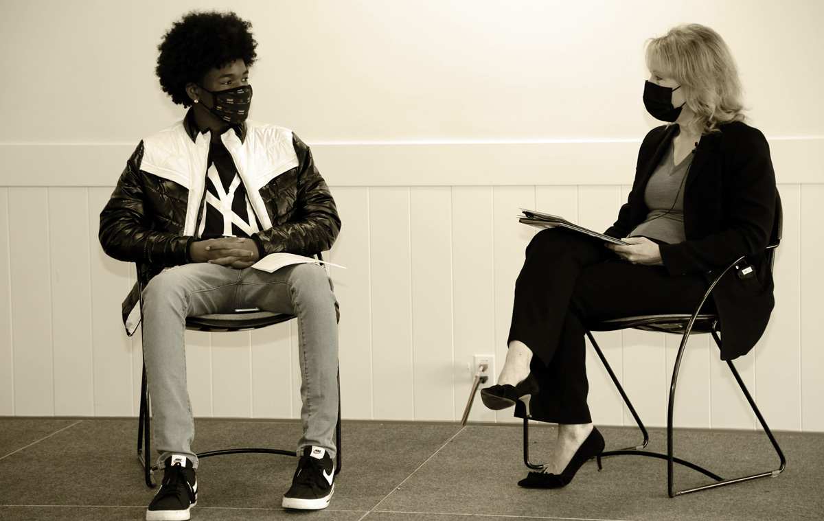 a masked woman and young man sit in chairs during a discussion