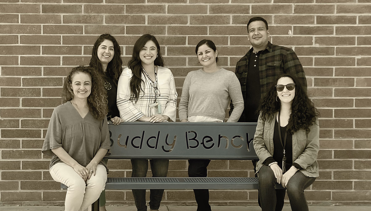 Group of people posing on a bench with the words "Buddy Bench" on it