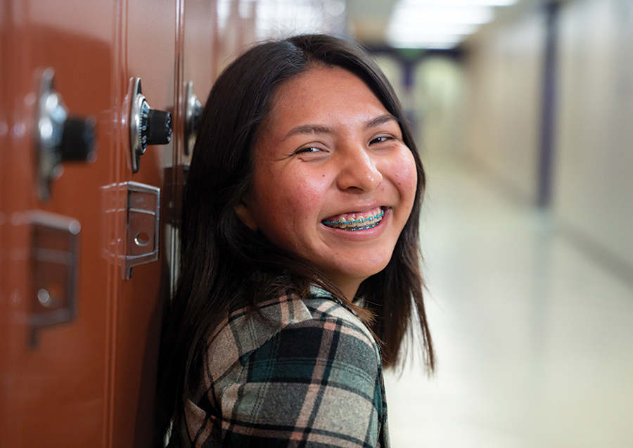 Student smiling and leaning against a red locker in the hallway of a school