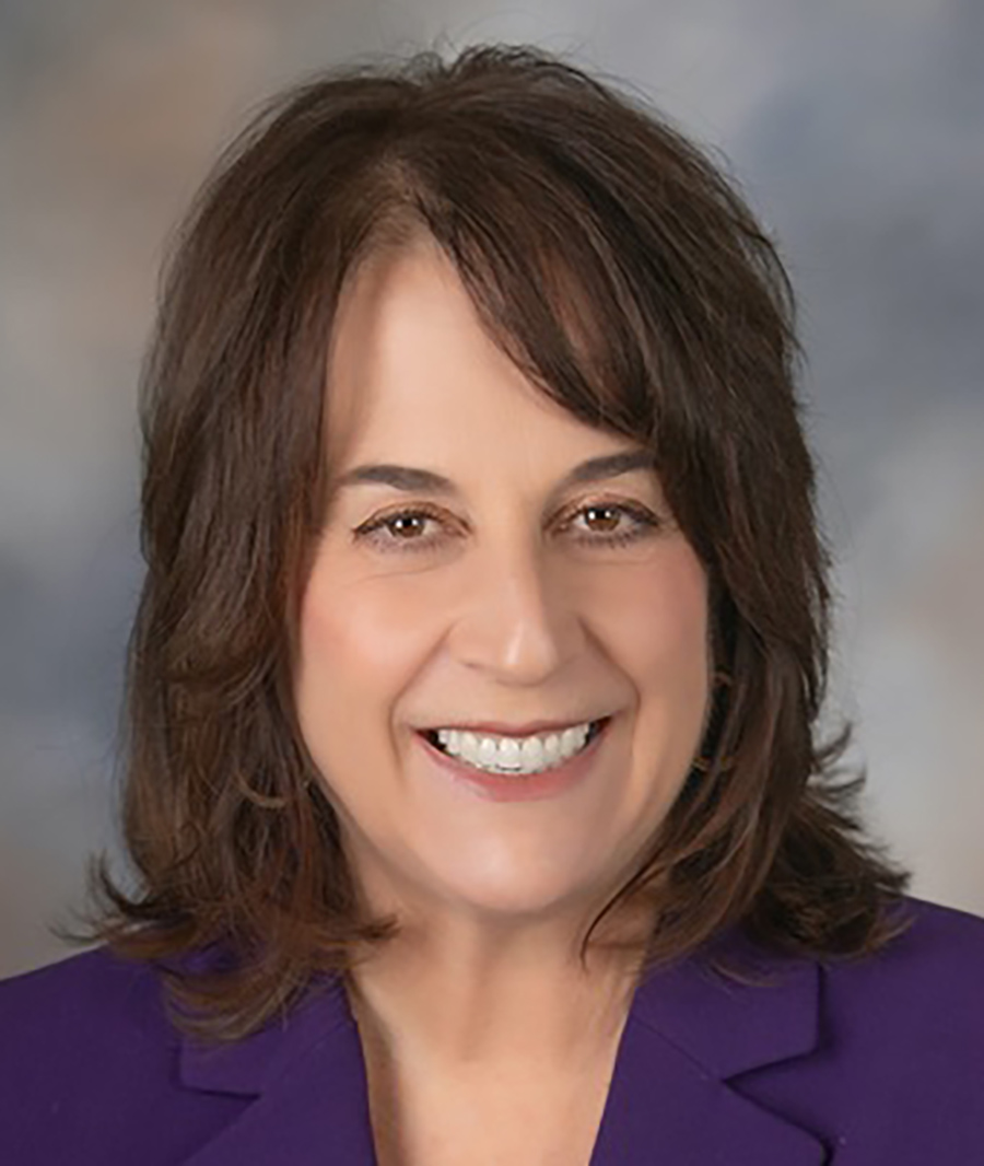 A portrait headshot photograph of Gina Cuclis smiling in a purple blouse