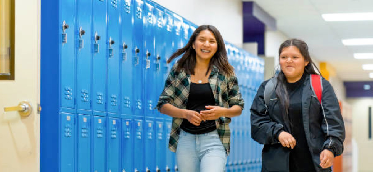 two students walking past blue lockers in the hallway while wearing their backpacks