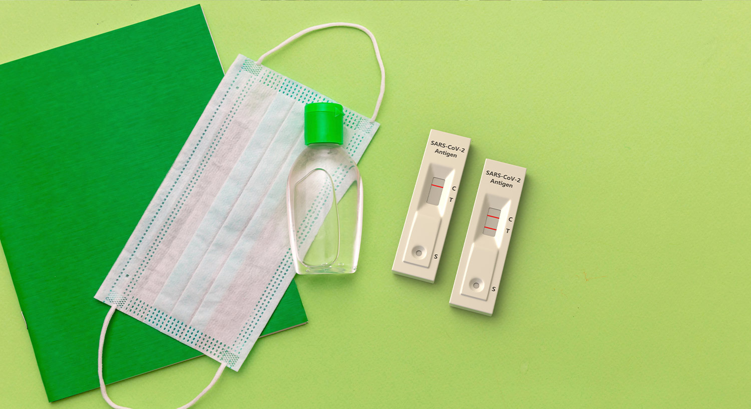 a face mask, hand sanitizer and two COVID tests on a green background