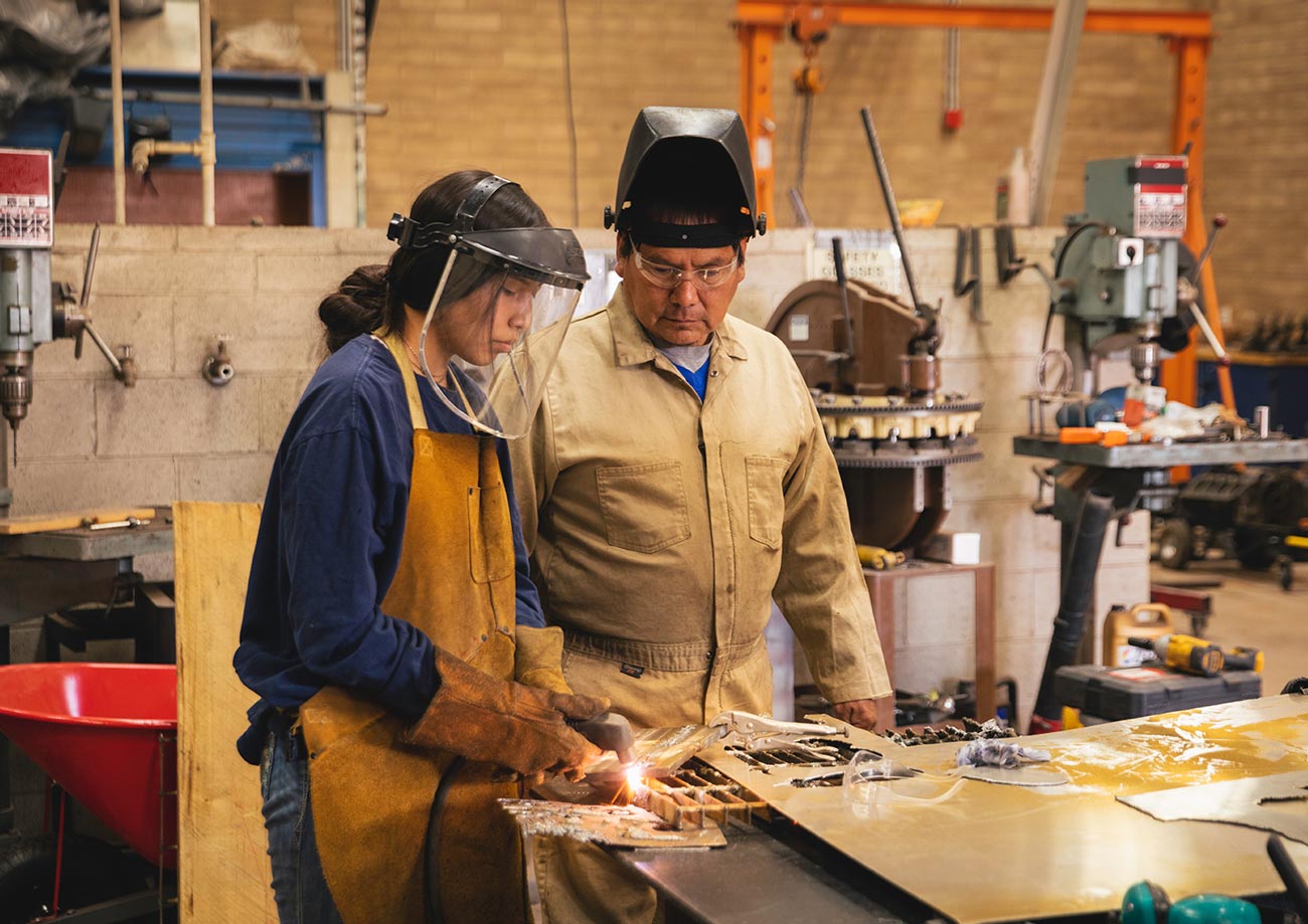 a teenaged student is supervised by an adult man while working with a welding tool in a shop class