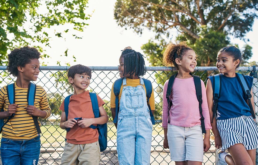five elementary aged children wearing backpacks smile at each other while standing against a chain link fence