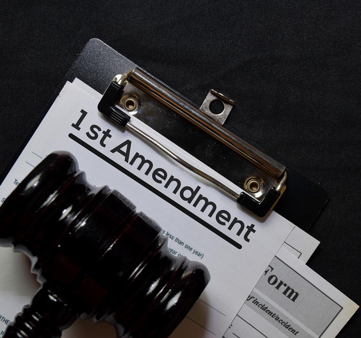 a judges gavel sits on a clipboard with the top slip of paper reading "1st Amendment"