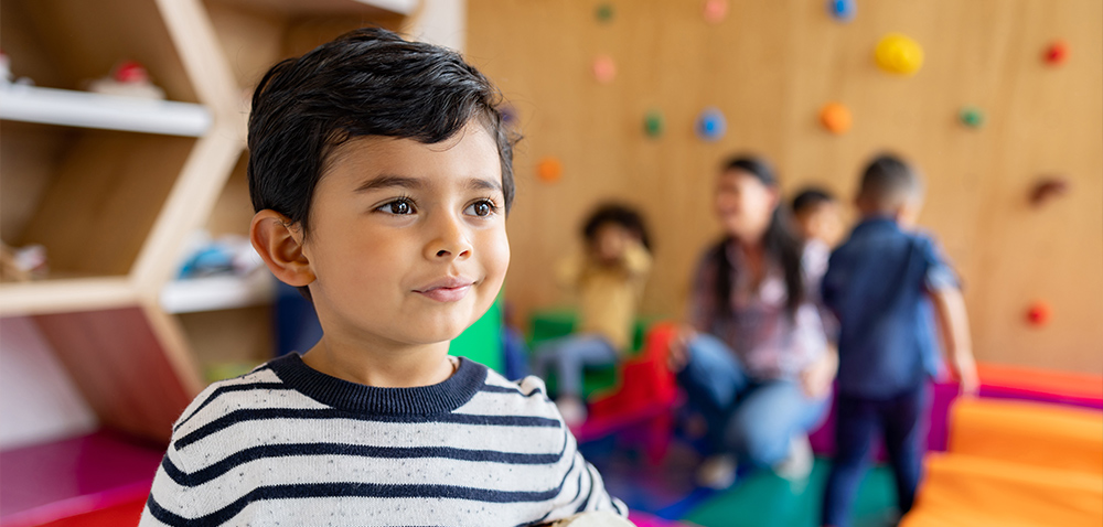 a kindergarten aged little boy stands with a slight smile in the foreground while a teacher and other classmates play out of focus in the background