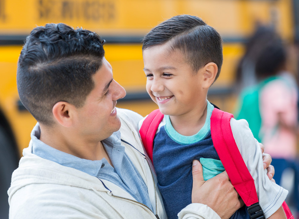 a father hugs his smiling elementary aged son with a school bus and children out of focus in the background