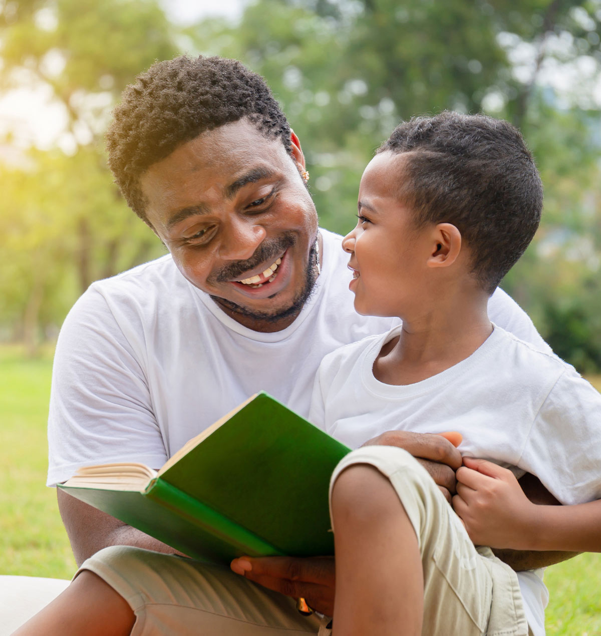 a young boy sits on his fathers lap outdoors while the two read a book together