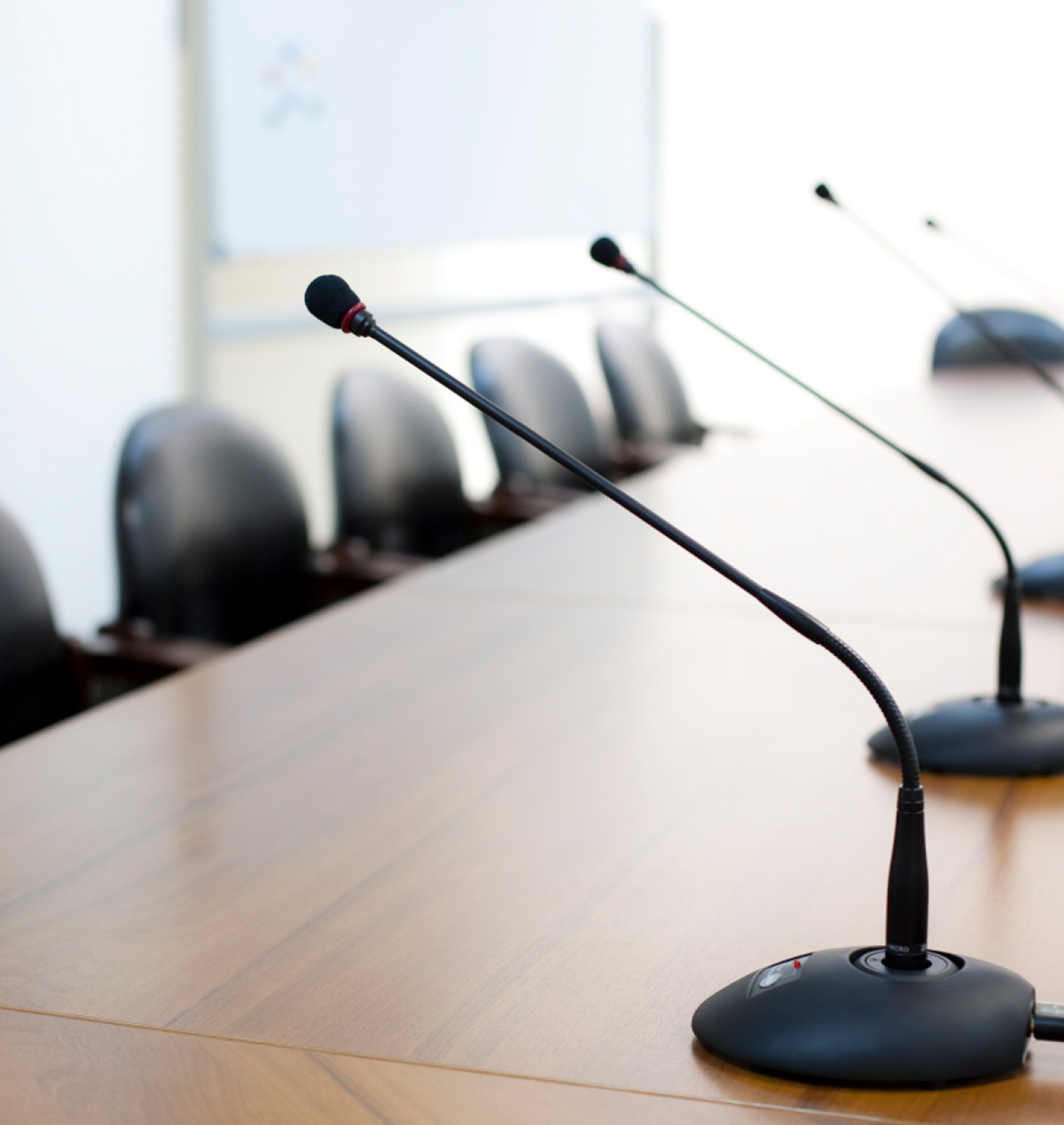 close up of a conference room table with a row of chairs behind and microphones atop