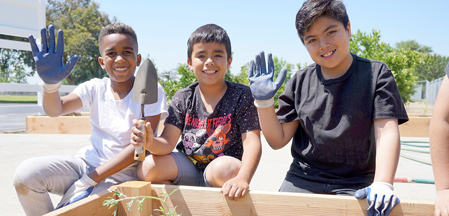 three young boys smile and wave at the camera while kneeling in front of a raised wooden planter