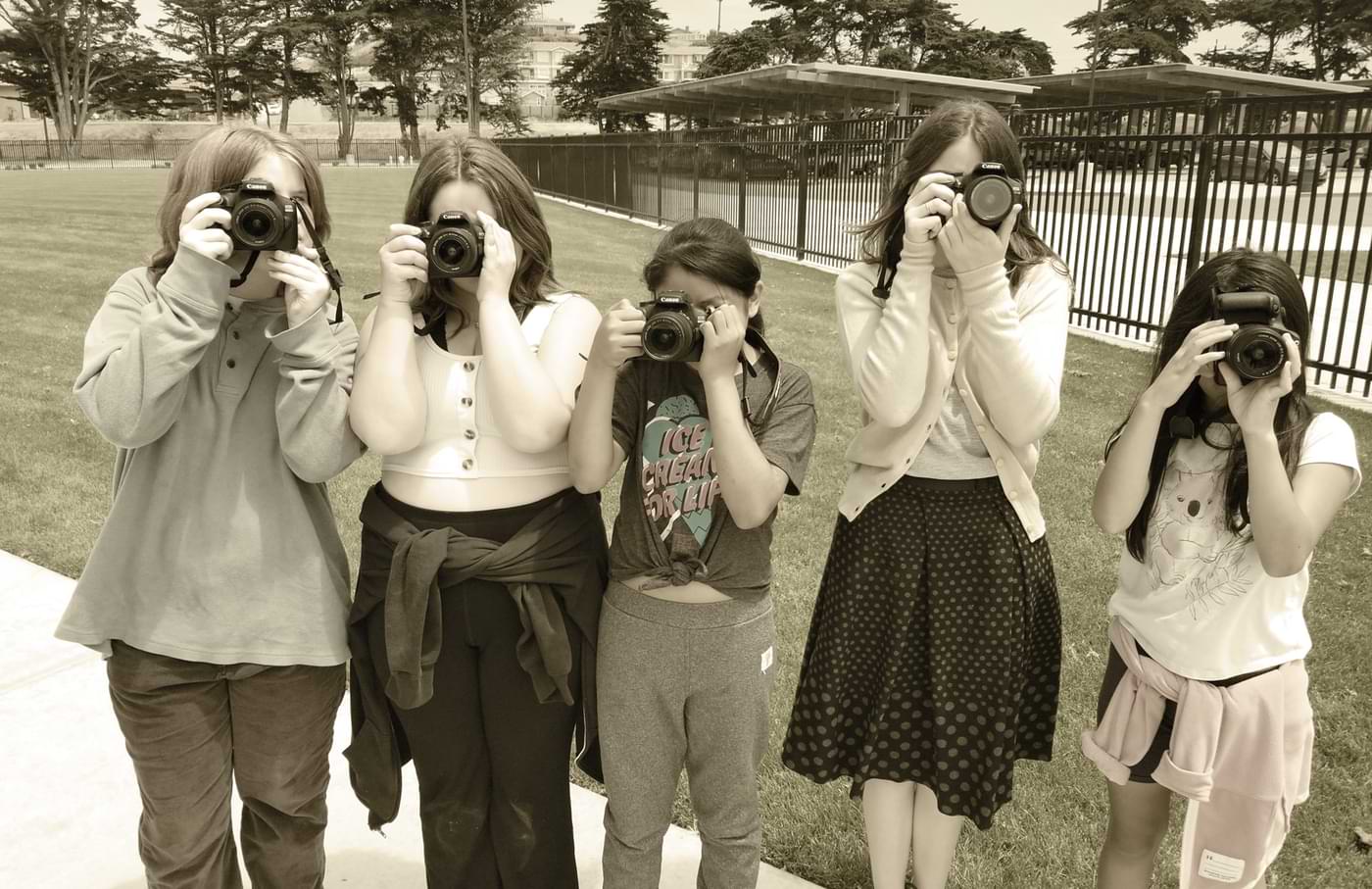 front view of five young girls standing in a schoolyard, each holding a canon camera to their eye
