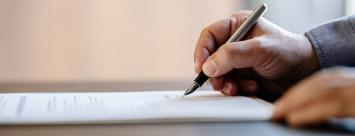 Close-up landscape photograph perspective of a person's right hand using a pen to sign on top of a white paper document