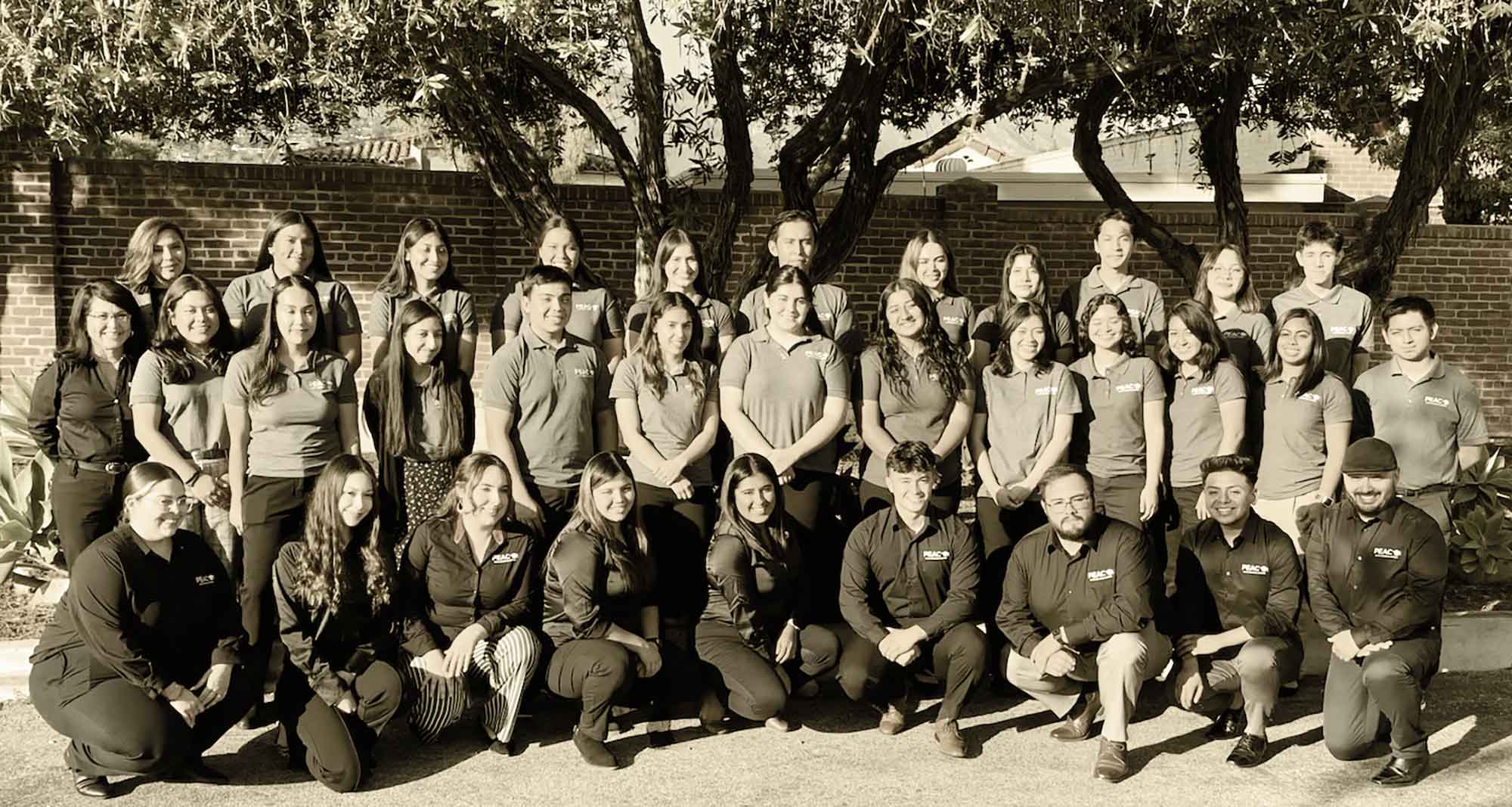 Landscape photograph of some Santa Barbara Unified teachers, administrators, and students smiling and posing together for a picture outside nearby a few trees and a brick wall