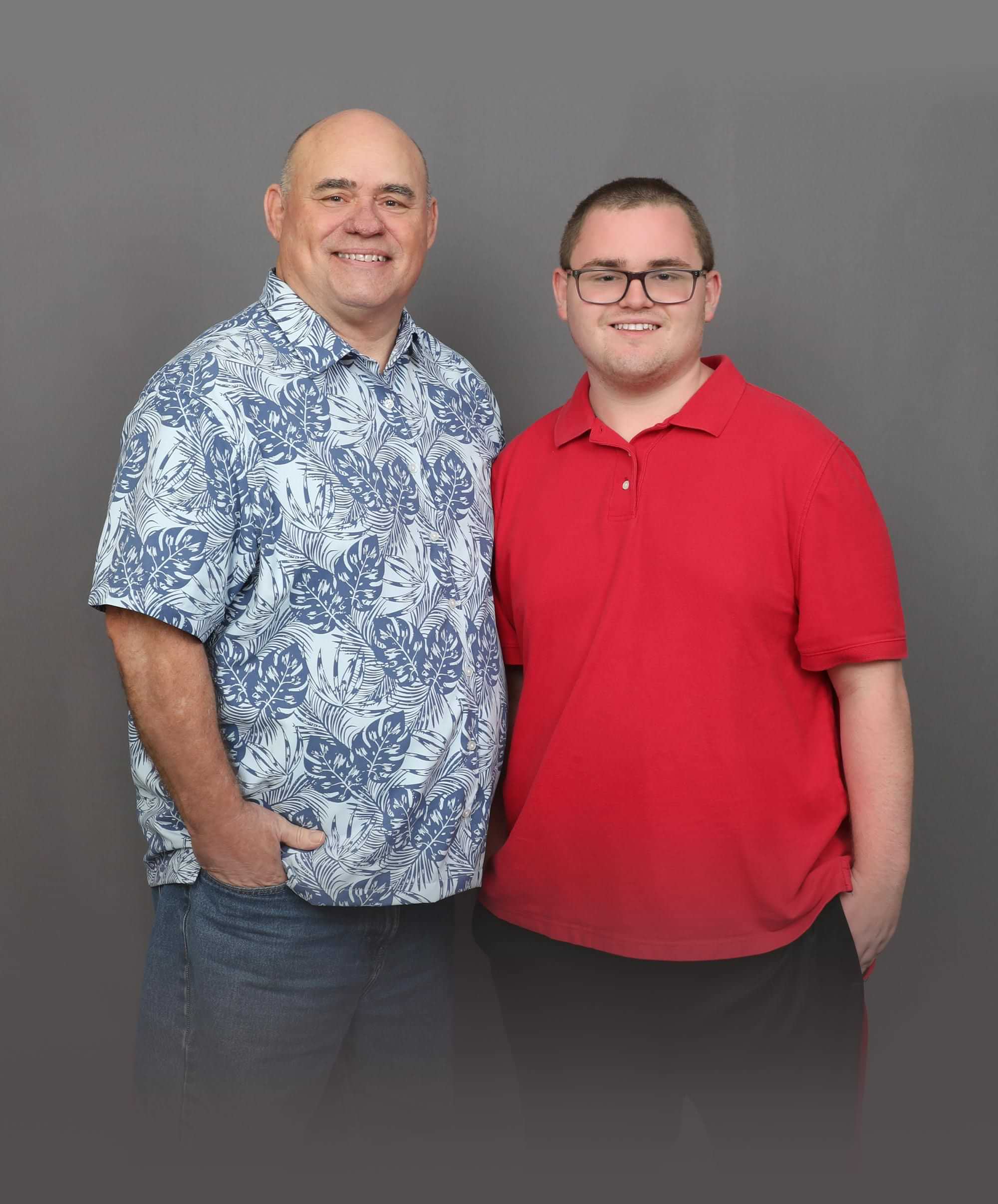 Robert Brown wears a blue monstera patterned short sleeve button down shirt and smiles while standing beside his smiling son, Joshua, who wears thick rimmed glasses and a red polo shirt