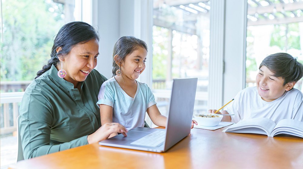 a woman of Native Amercian descent smile and sits at a table on a computer while a young girl smiles while sitting on her lap and a young boy does work on another end of the table