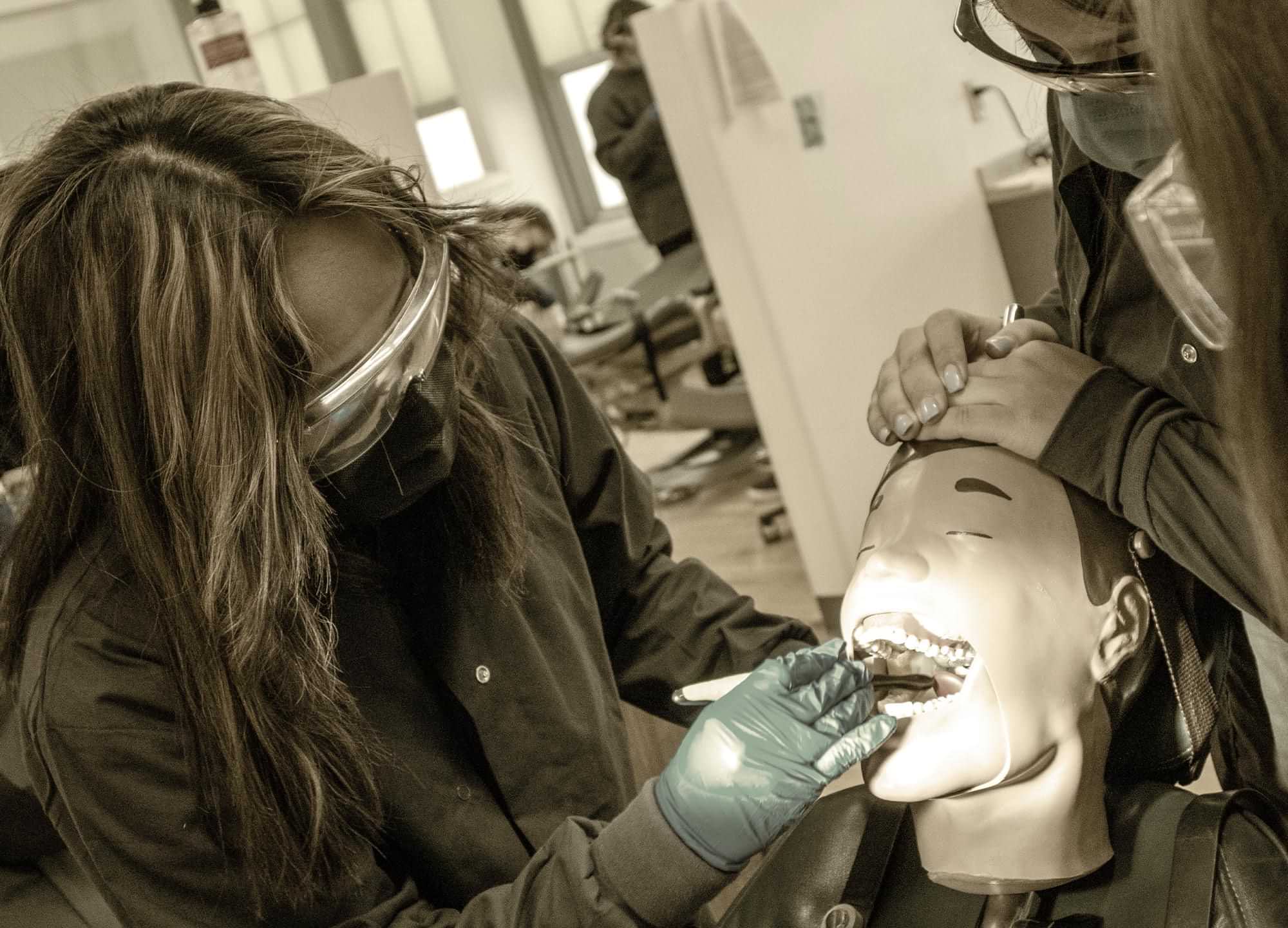 students wearing masks and goggles perform mock dental work on a realistic dental mannequin head