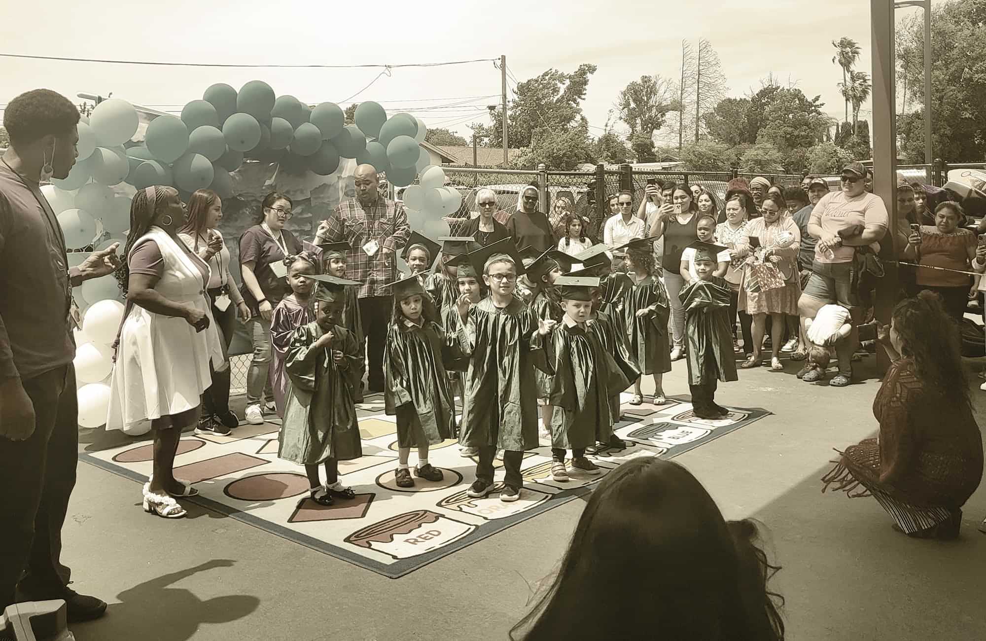 young students of the Mary Bird Early Childhood Education Center dressed in caps and gowns stand on a colorful shape rug in the center of a large crowd of parents, all gathered for a mini matriculation event