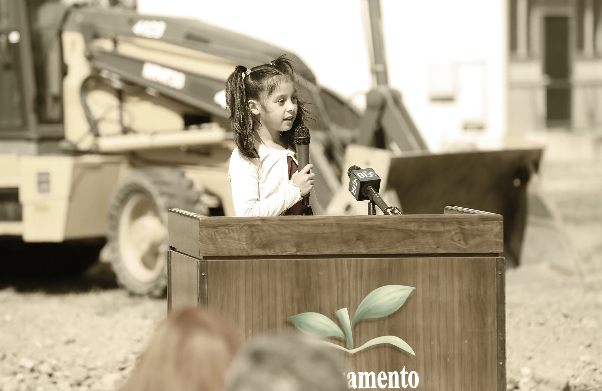 a young female Sacramento City Unified School District students stands outside at a podium, speaking into a microphone on a bright day