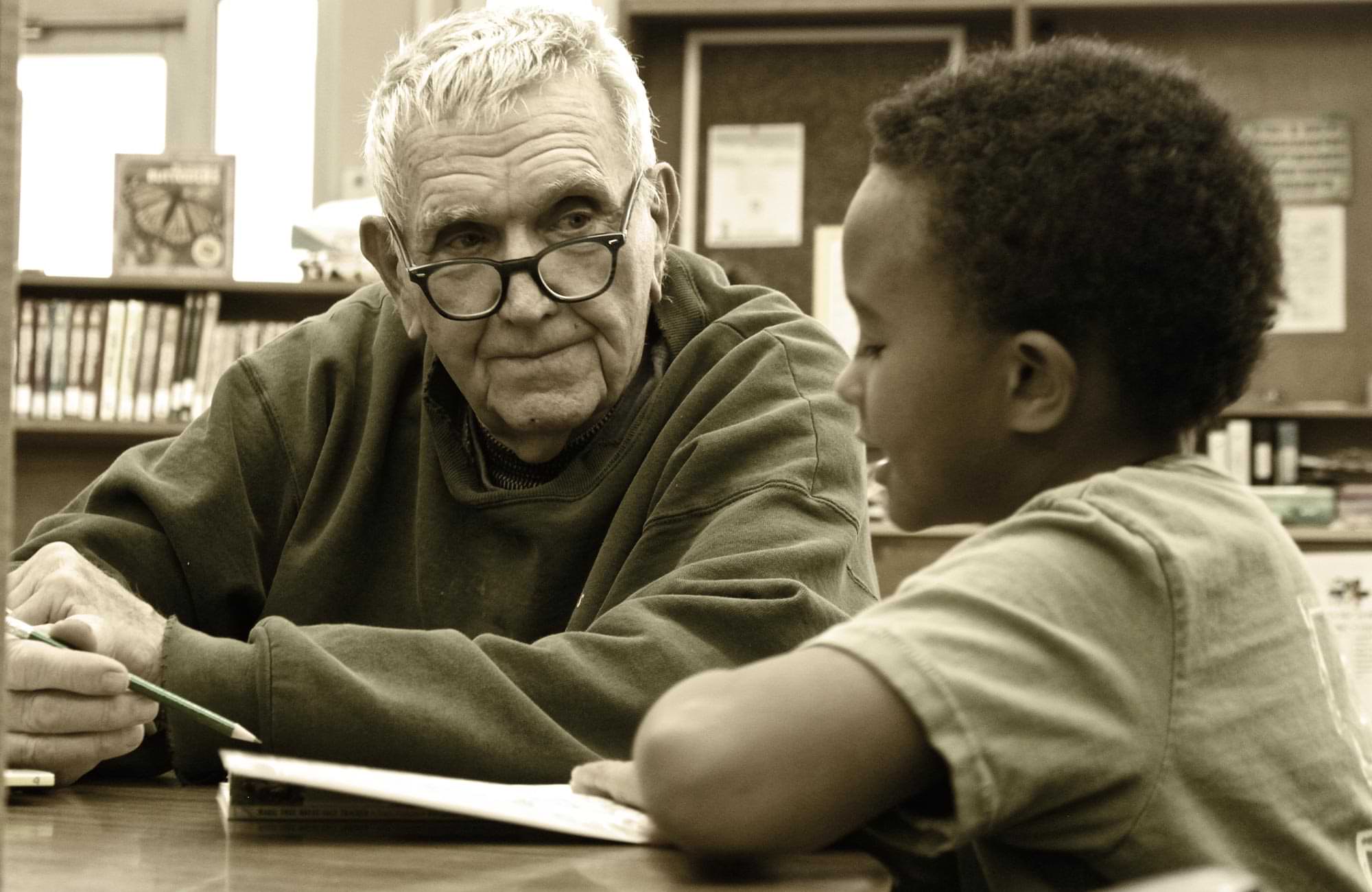 an older gentleman watches and guides with a pencil as a young boy sits beside him and reads