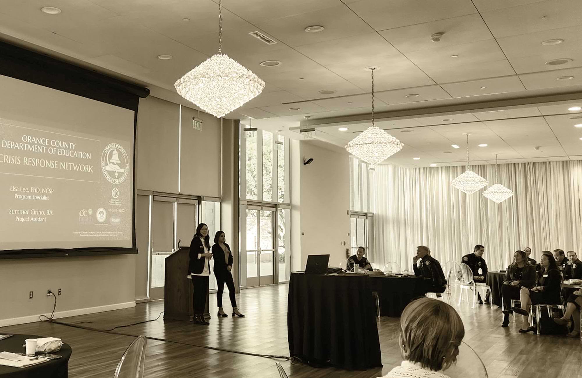 two women stand by a podium addressing a large audience at a Crisis Response Network meeting