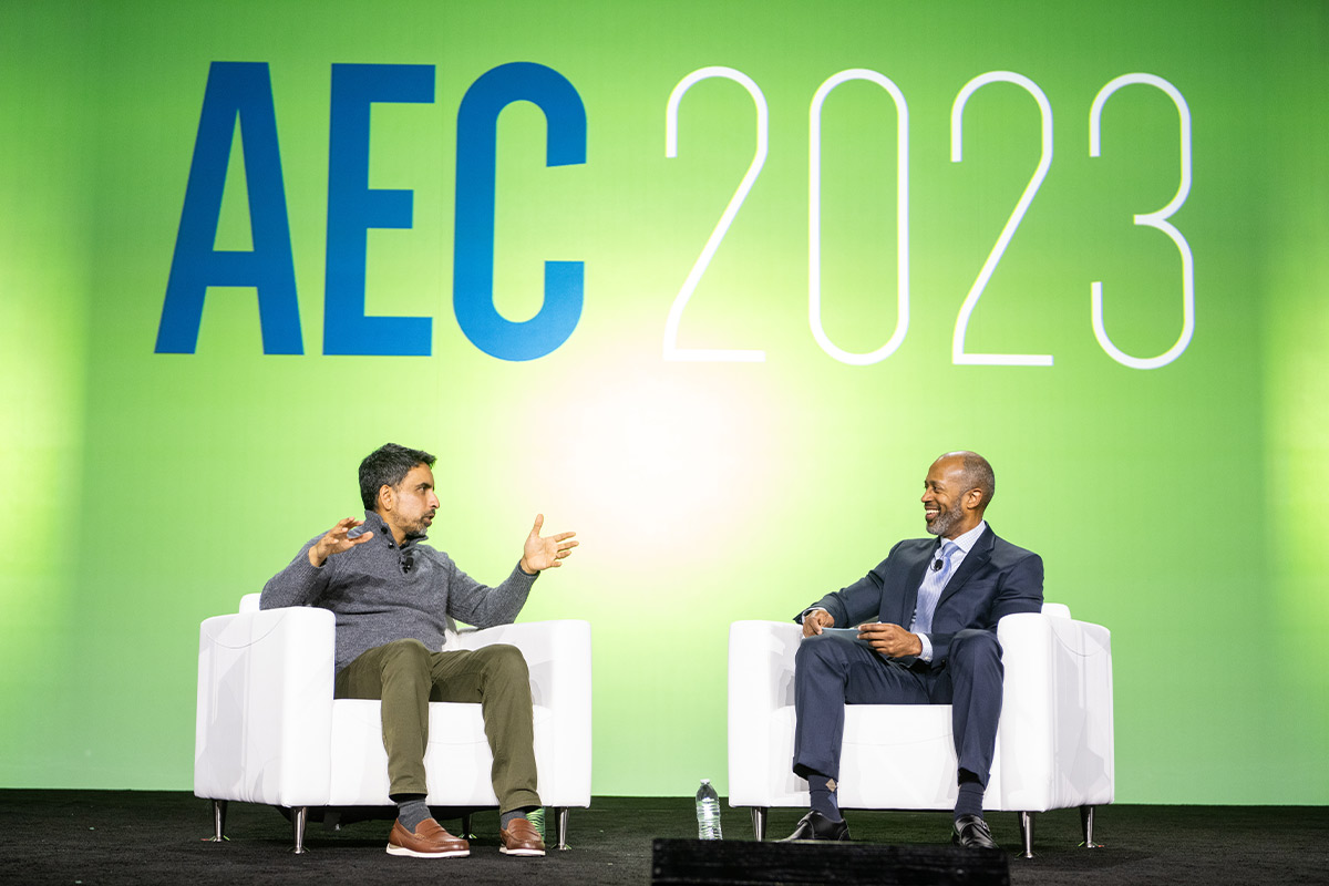 Sal Khan and Vernon M. Billy each sit in a white armchair looking at each other in discussion while on a stage, the wall behind them is a bright lime green and displays the text AEC 2023
