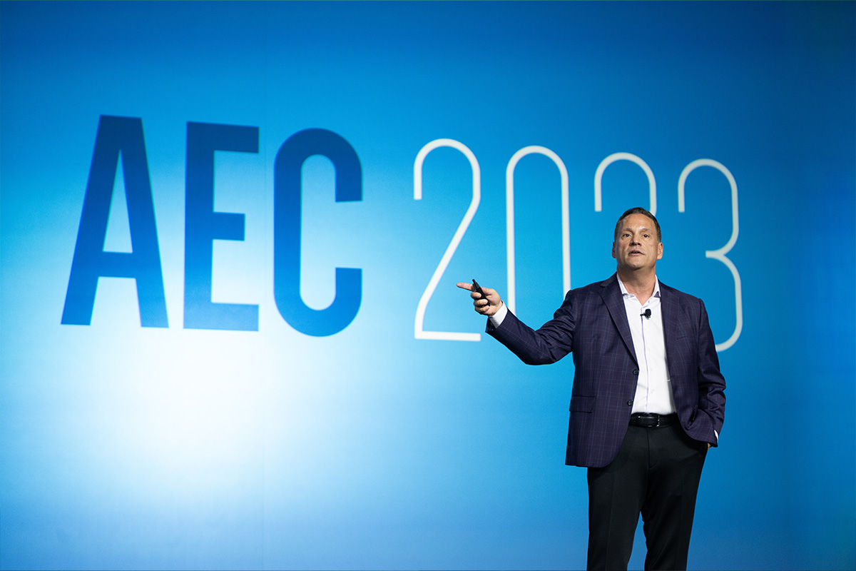 Mark C. Perna stands on on a stage wearing a blue plaid jacket and black slacks, the wall behind him is a bright blue green and displays the text AEC 2023