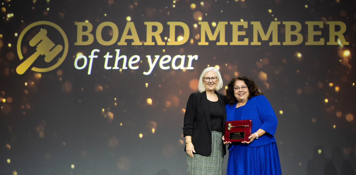 CSBA President Susan Markarian stands on stage with the Golden Gavel winner, Mary Helen Ybarra from Corono-Norco USD; Mary Helen Ybarra holds her plaque as both women stand against a large screen that reads: Board Member of the Year