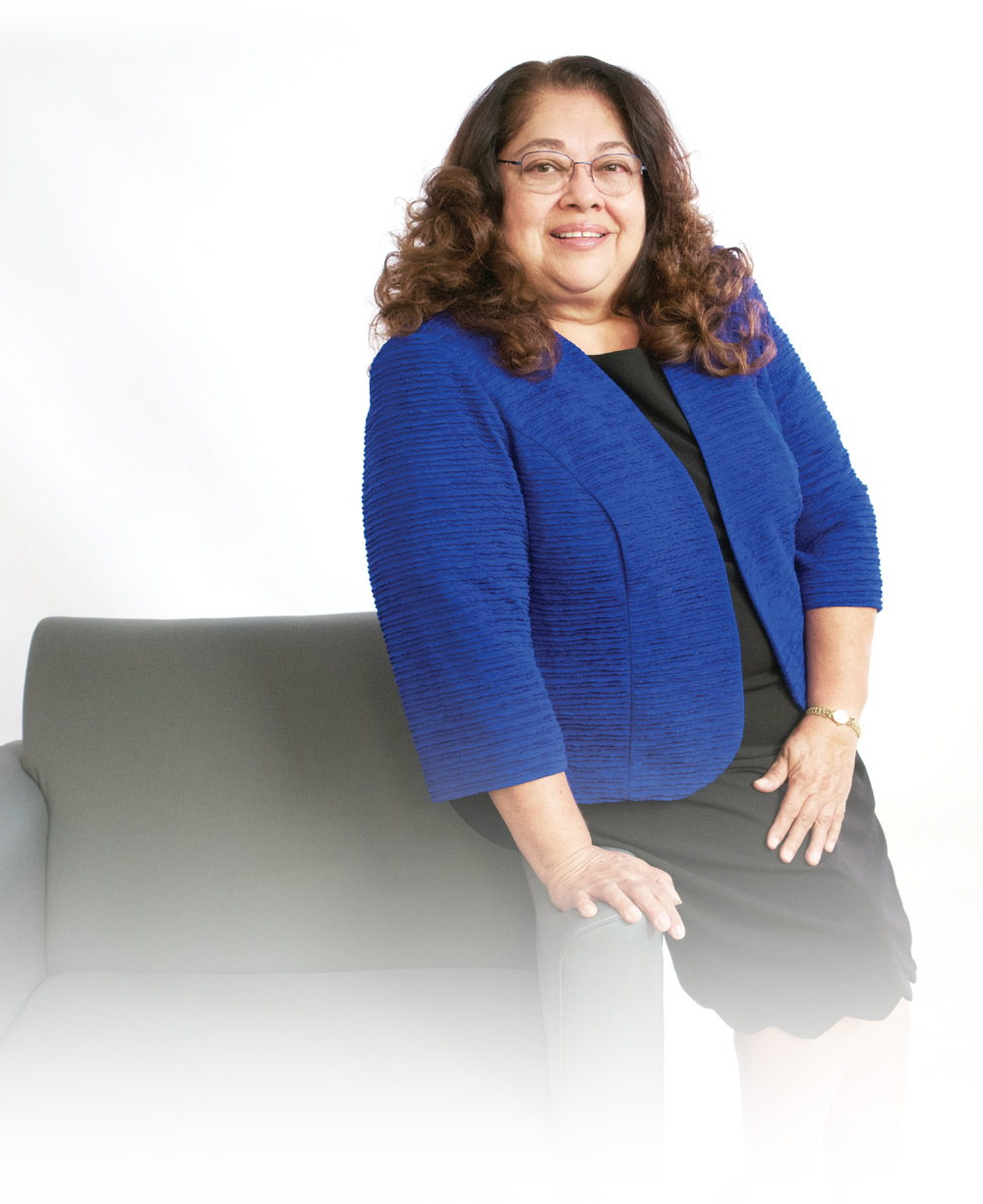 Mary Helen Ybarra leaning against a chair while wearing a blue blazer and black dress