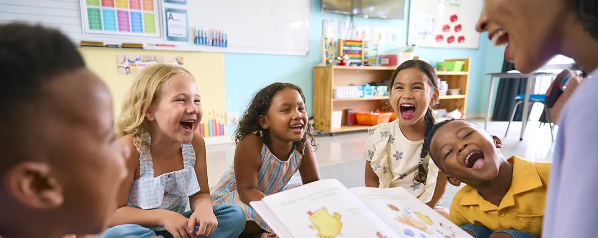 Landscape close-up photograph indoor school classroom view of a teacher reading a children's book to her students as they all smile in joy