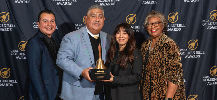 four people standing together while the two people in the middle hold an award together in front of a golden bell awards background 