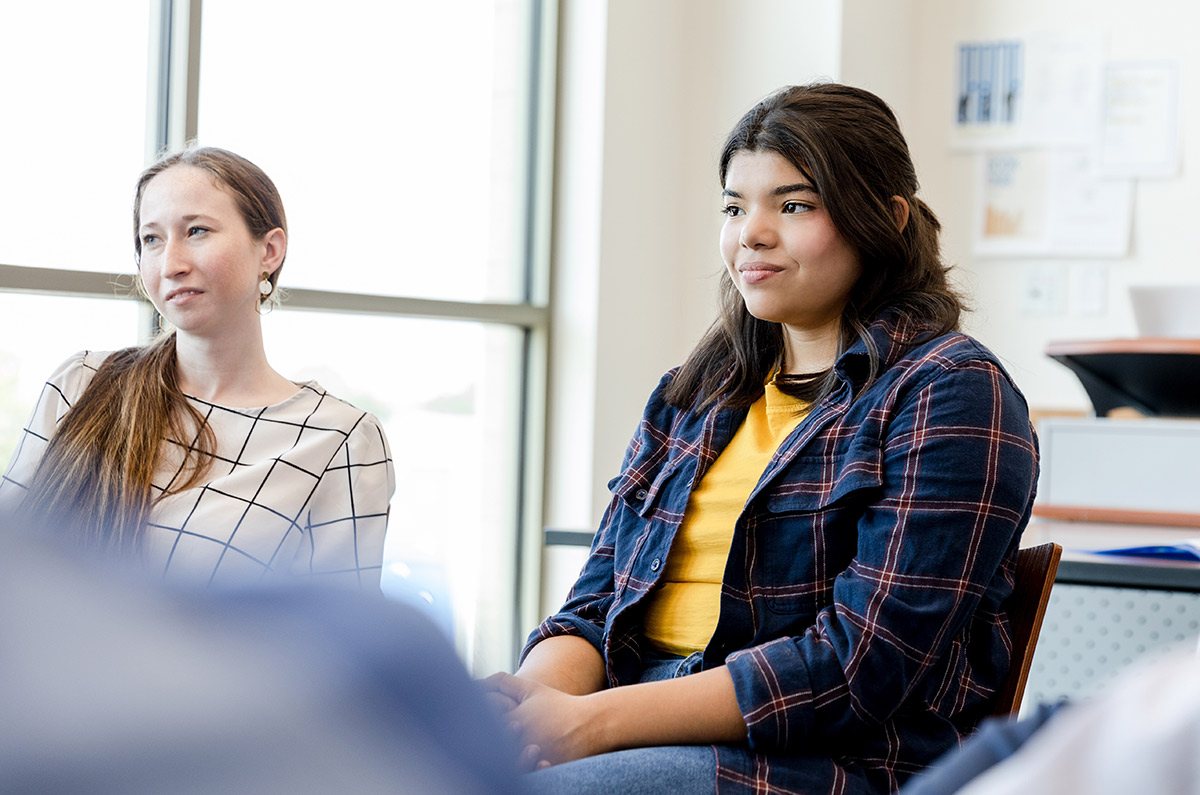 Landscape close-up photograph view of two young female individuals seated down in chairs amongst a mental health discussion group inside an open-area office room of some sort