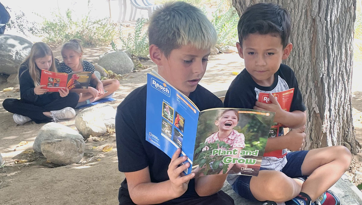 Two young boys reading a book together surrounded by nature and other students