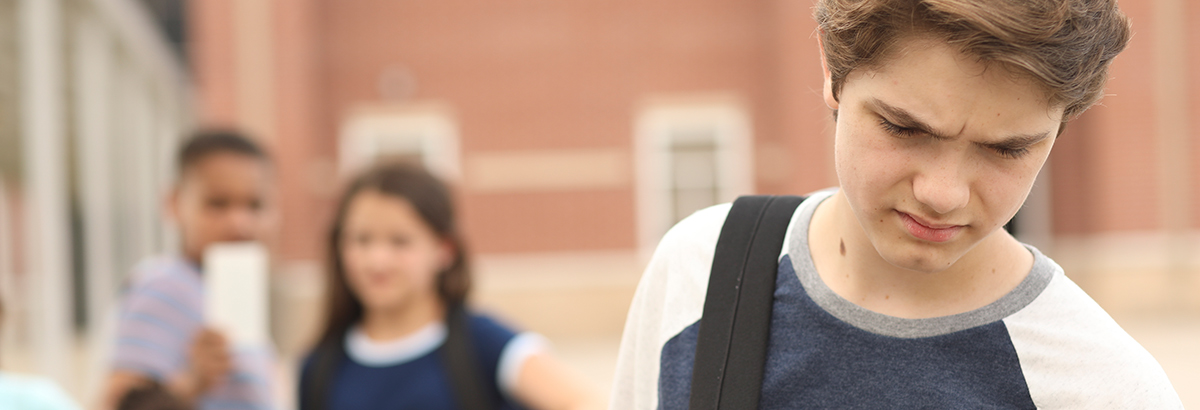 Landscape close-up photograph cropped view of a young boy carrying a backpack as he has his head down looking frustrated as there are two other young kids (one boy and one girl) in the background watching him walk away