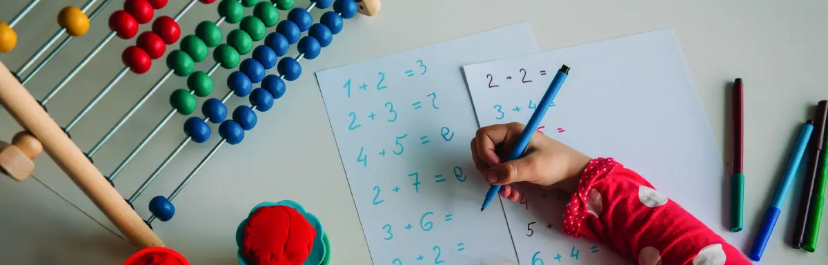Landscape orientation close-up photograph cropped top aerial view of a little young kid using a thin blue colored marker to write down some math equation numbers as in front of this individual are different school supplies/materials in what appears to be a abacus mathematical calculating object, red circular shaped play-doh, other various thin colored markers, and two white sheets of paper that contain numerical math equations on them such as 1 + 2 = 3, etc.