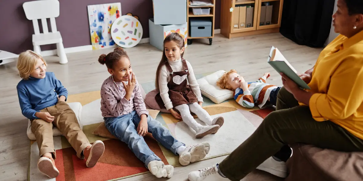 Landscape orientation close-up photograph view of a female teacher reading a book to four young little kids inside a classroom as everyone is seated down on the floor and one of the four young little kids is a female individual smiling/laughing by touching at her own nose with her own fingers