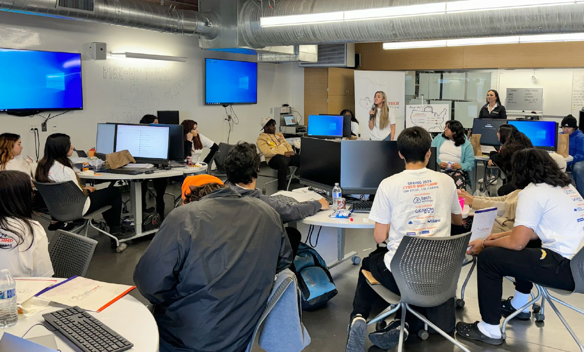 students in a computer lab focused on a woman speaking into a microphone at the front of the room