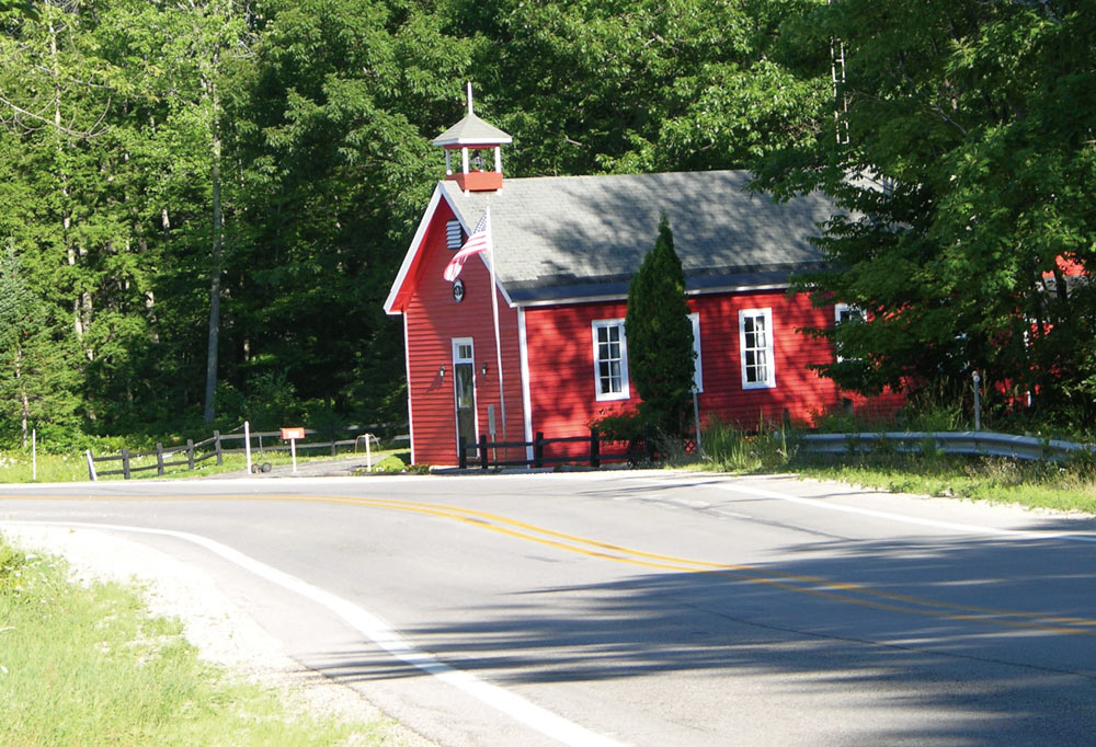 A small red building with a gabled roof and belfry by a curved road, surrounded by green trees.