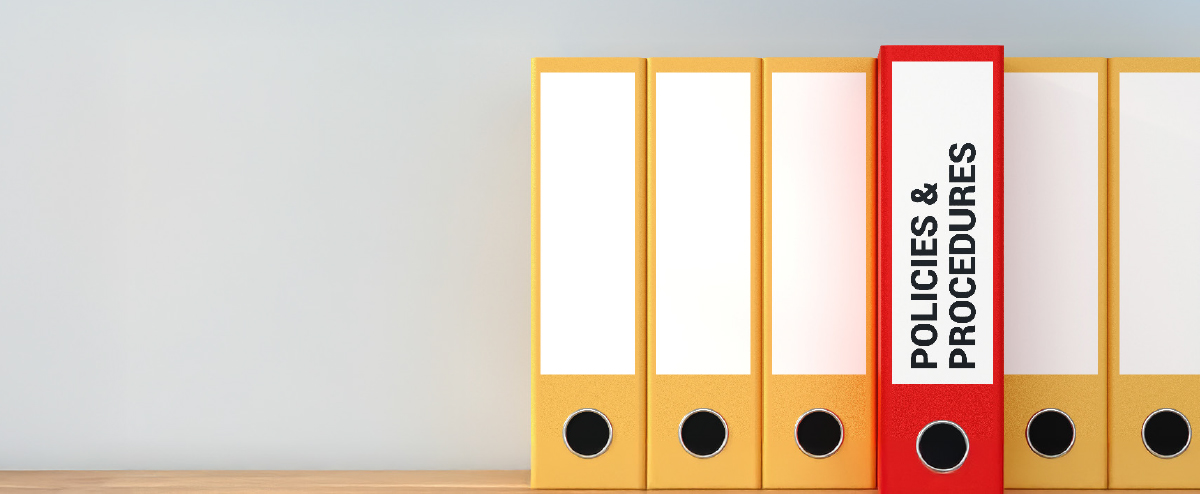 Digital landscape orientation cropped image view of a row of yellow file binders with one red binder labeled POLICIES & PROCEDURES standing out, against a light gray wall and light wood surface