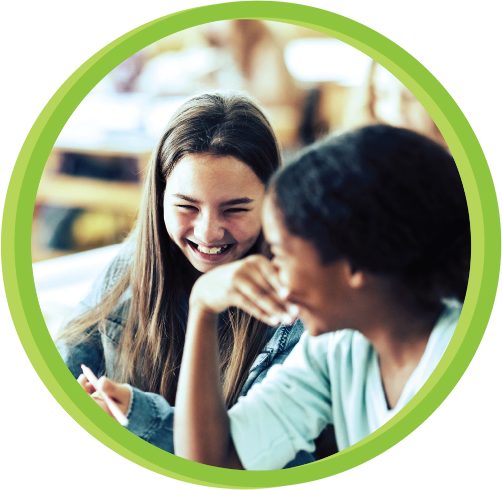 circular crop image of two young girls laughing while sitting beside each other in a classroom