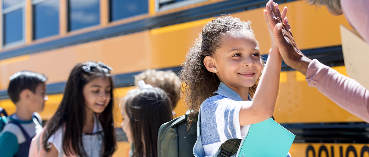 students in a line on the side of a school bus high-fiving a teacher