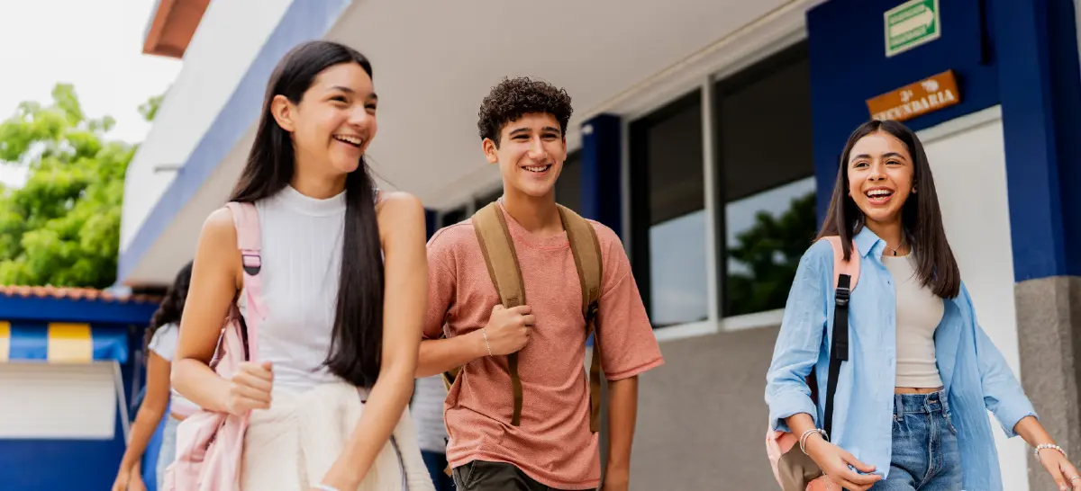 Cropped digital stock photograph image of three smiling teenagers each wearing a backpack walking outdoors alongside a modern school building on campus grounds; The girl on the left has long dark black hair and wears a light top and pink backpack; The boy in the middle has black curly hair and wears a dark faded pink t-shirt and tan backpack; The girl on the right has long dark black hair and wears a light blue open button-down dress shirt, off-white top, and pink backpack