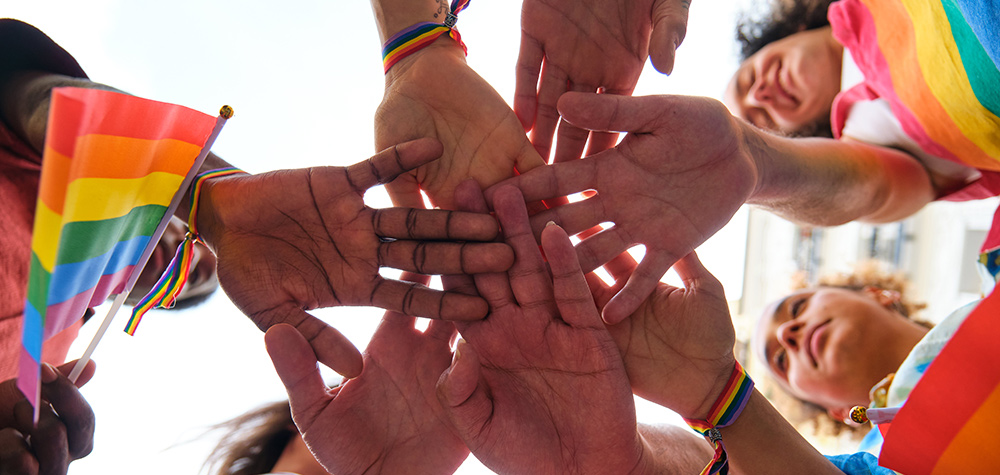 A diverse group of people put their hands together in the center while holding small rainbow Pride flags.